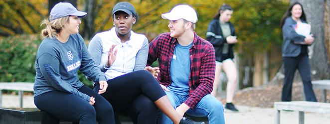 Three students with baseball caps, seated in discussion on benches